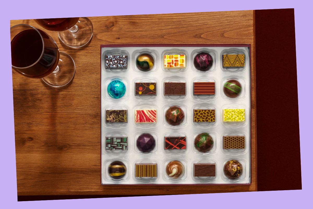 A box of luxury chocolates on a wooden table next to two glasses of red wine