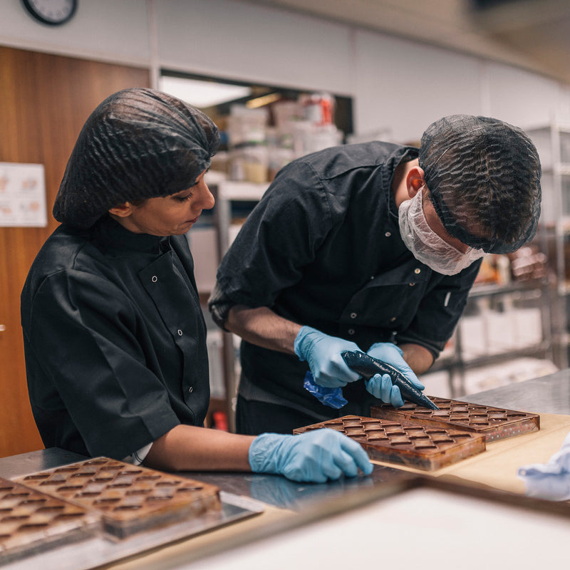 A young male chocolatier being supervised as he fills chocolate moulds