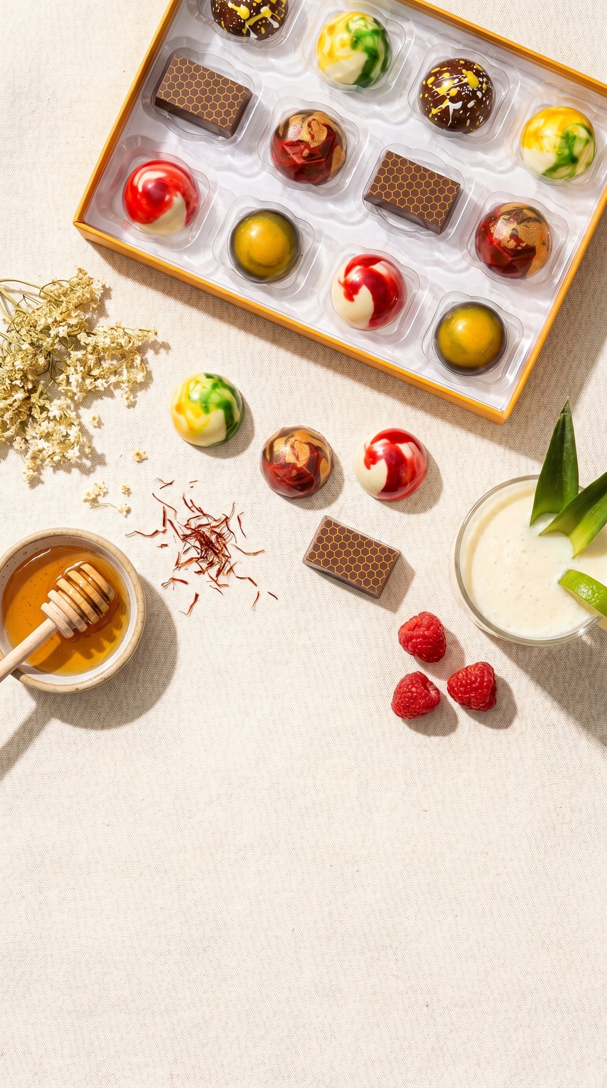 Assorted chocolates in a box with honey, raspberries, and a pina colada on a light surface.