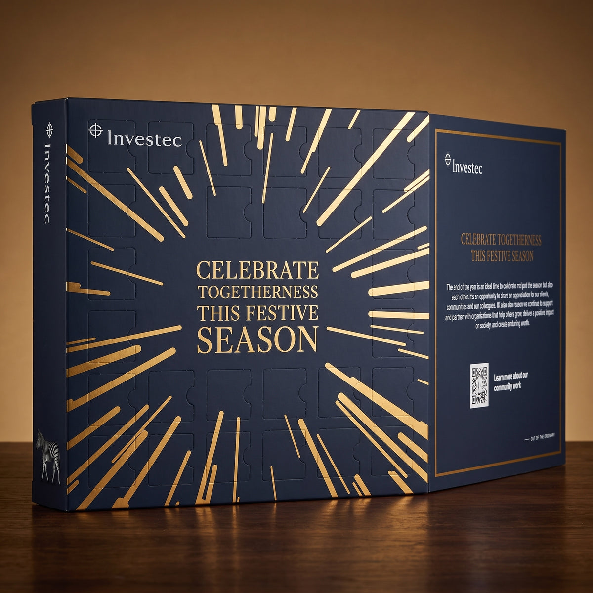 A dark navy blue Investec advent calendar with gold foil pattern and gold text reading 'CELEBRATE TOGETHERNESS THIS FESTIVE SEASON,' photographed on a wooden surface against a warm brown background.