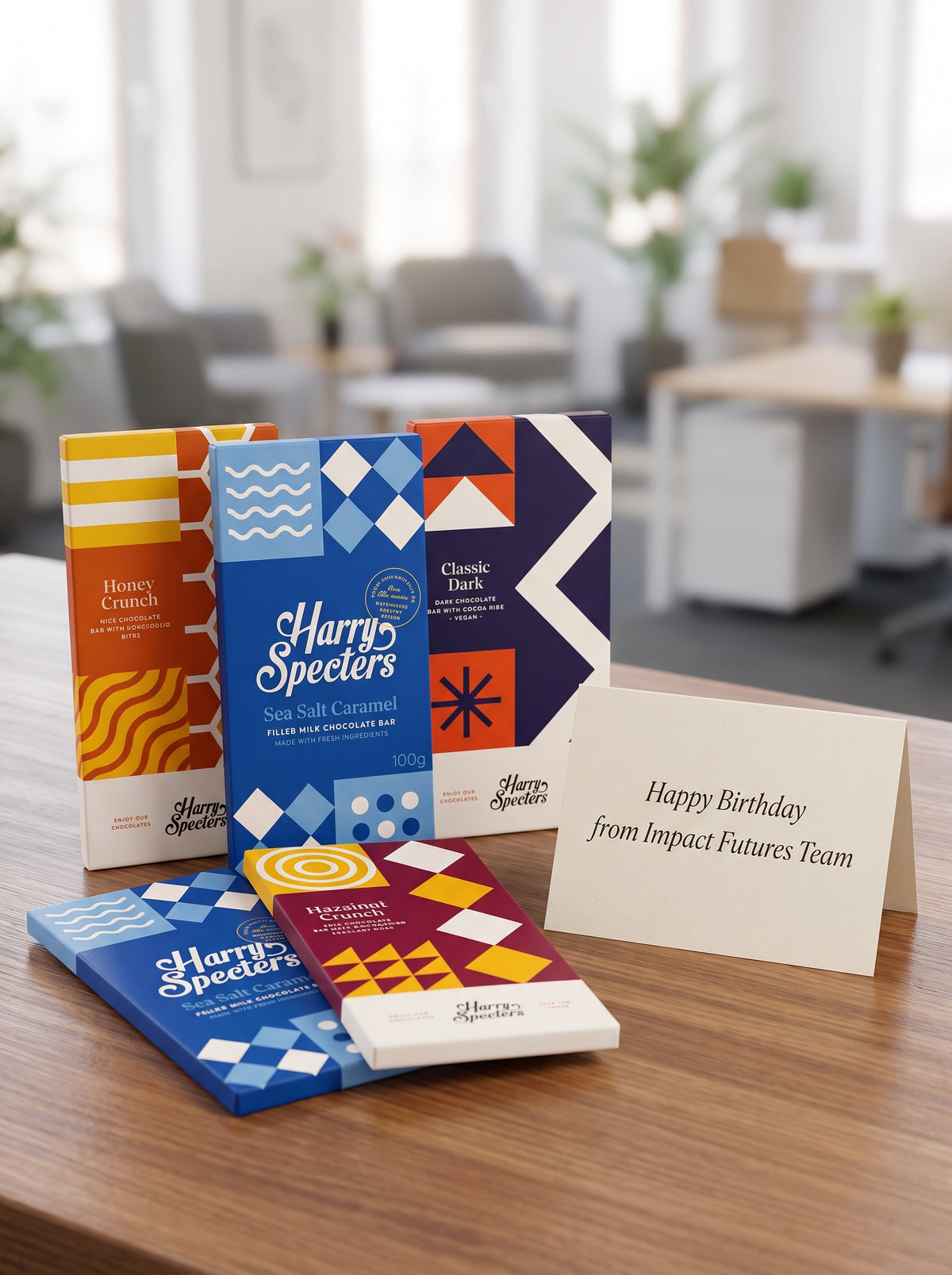 Three Harry Specters chocolate bars with colourful geometric packaging are arranged on a wooden table next to a white greeting card that reads 'Happy Birthday from Impact Futures Team.' The background shows a softly blurred office interior.