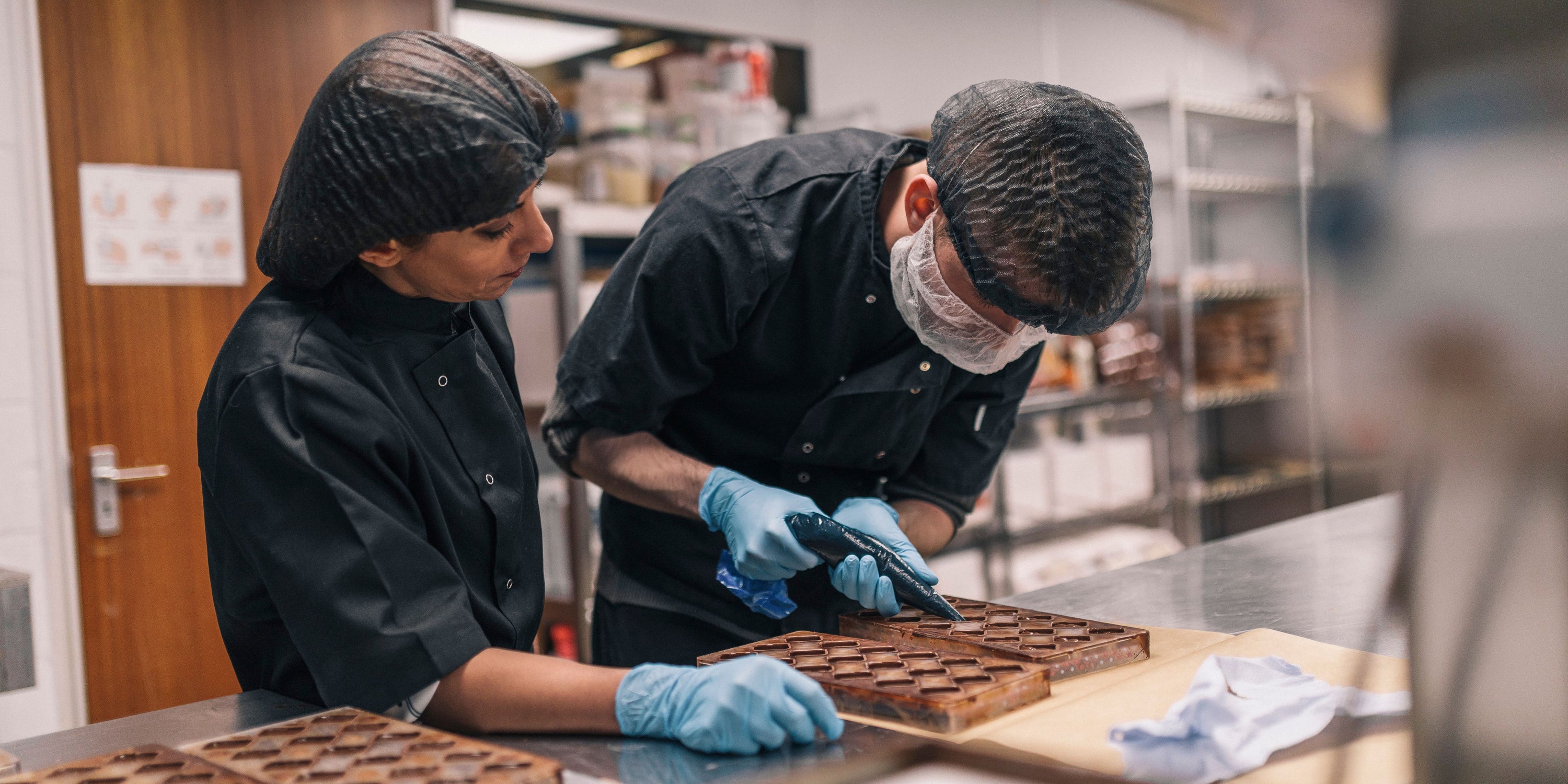 Two cholatiers working, one piping chocolate while the other observes.