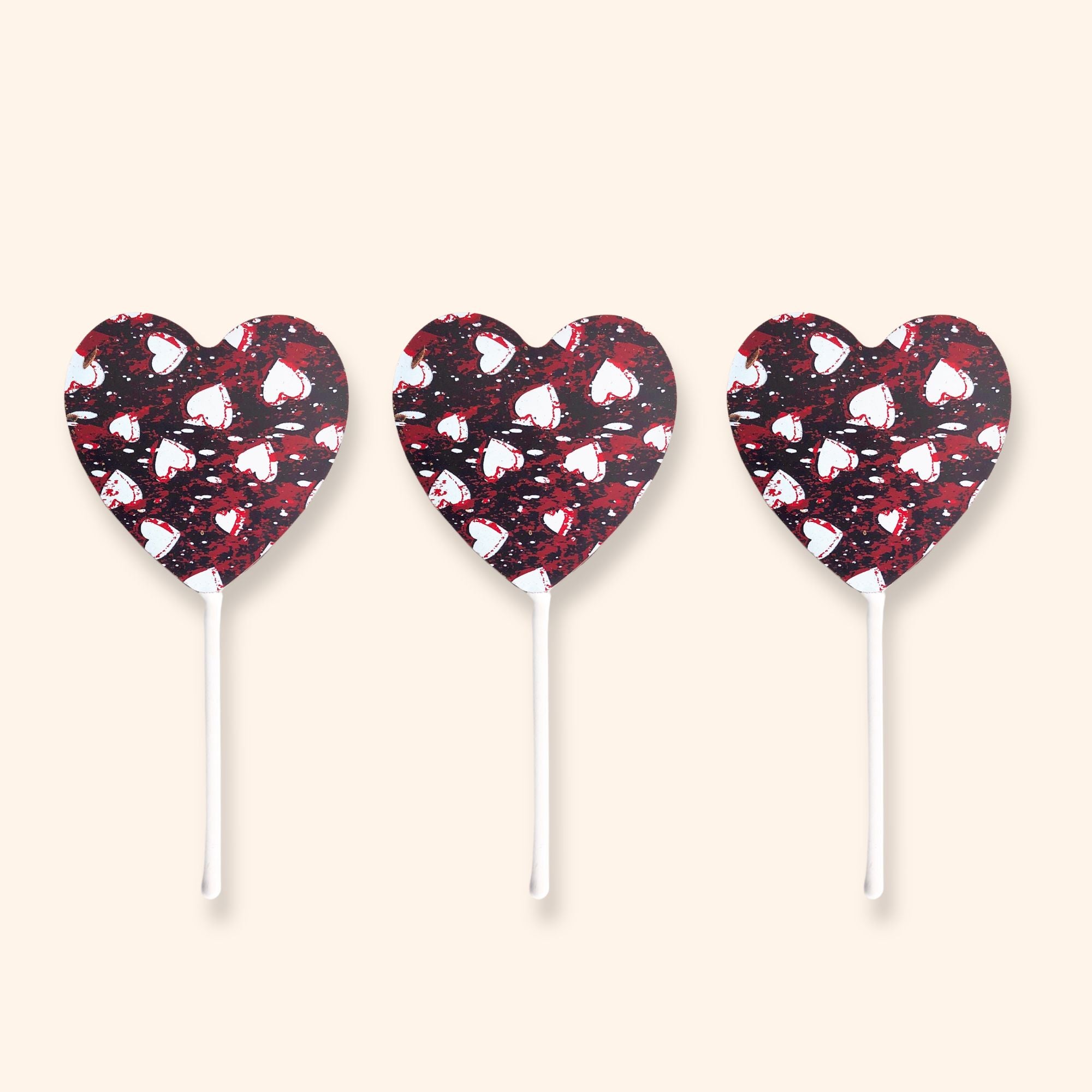 Three dark chocolate heart-shaped lollipops with a black and red pattern on a beige background
