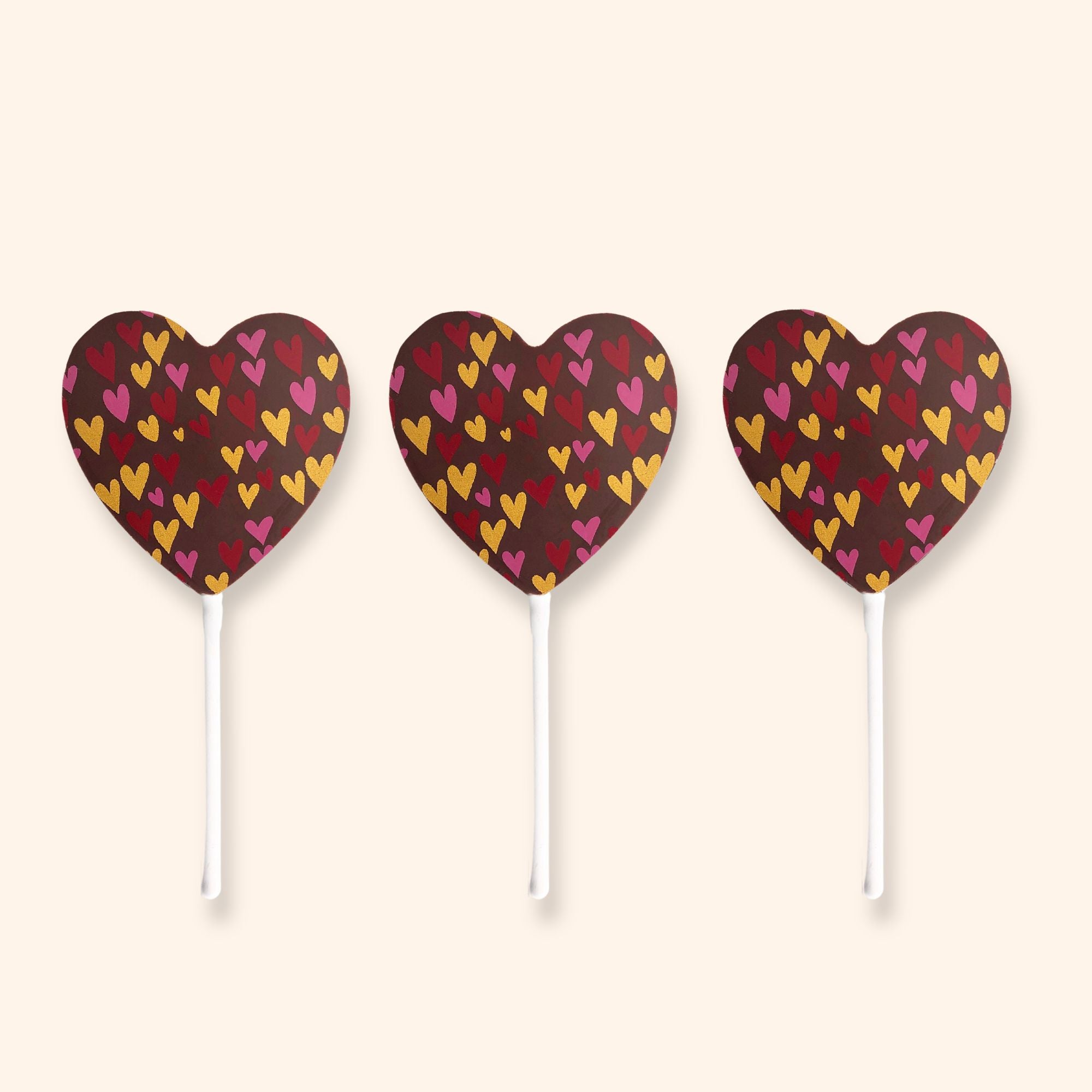 Three heart-shaped milk chocolate lollipops with colourful heart patterns on a beige background