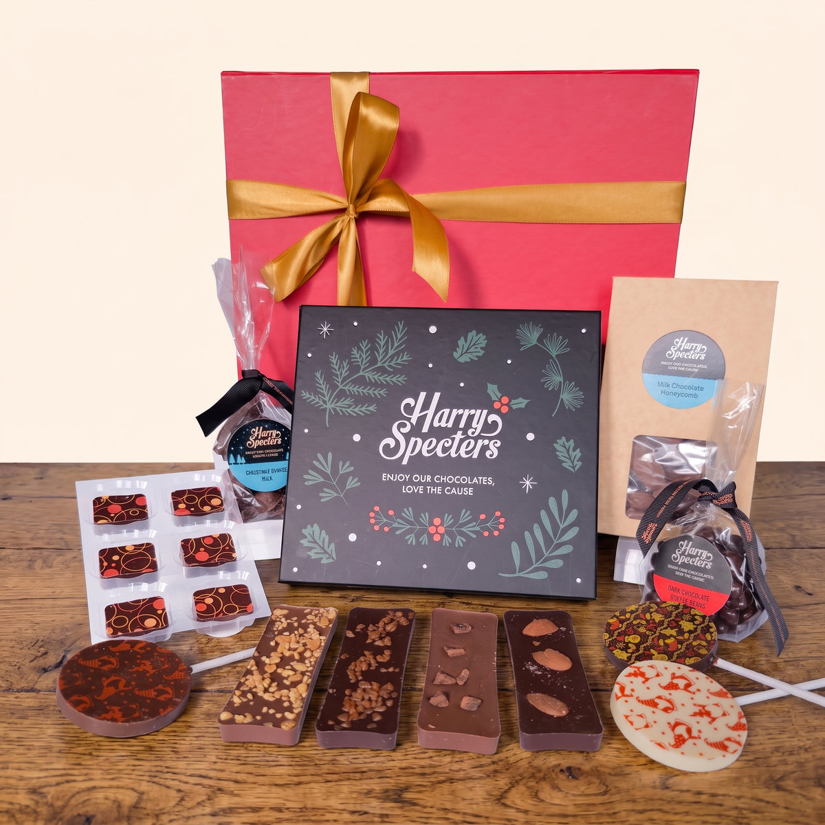 A chocolate gift set displayed on a wooden table with a large red gift box and gold ribbon behind it. In front is a black decorative box reading 'Harry Specters', along with assorted chocolates, lollipops, a bag of chocolate coffee beans, and a kraft paper bag of chocolate honeycomb.