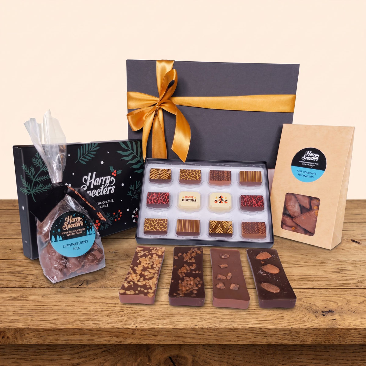 A chocolate gift set displayed on a wooden table against a warm beige background. The set includes an open box of assorted chocolates, a bag of chocolate shapes, a kraft paper bag of chocolate truffles, and a black gift box with green leaf graphics and a gold ribbon.