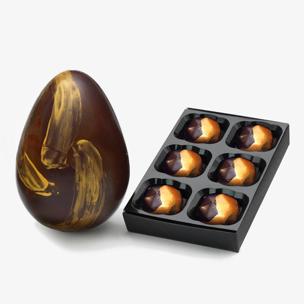 A dark chocolate Easter egg decorated with gold brush strokes next to a box of 6 dark chocolates