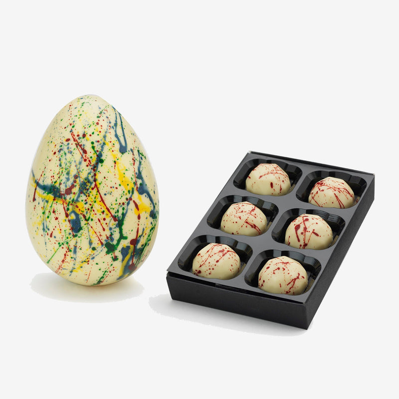 A white chocolate Easter egg decorated with rainbow splatter next to a box of 6 white chocolates